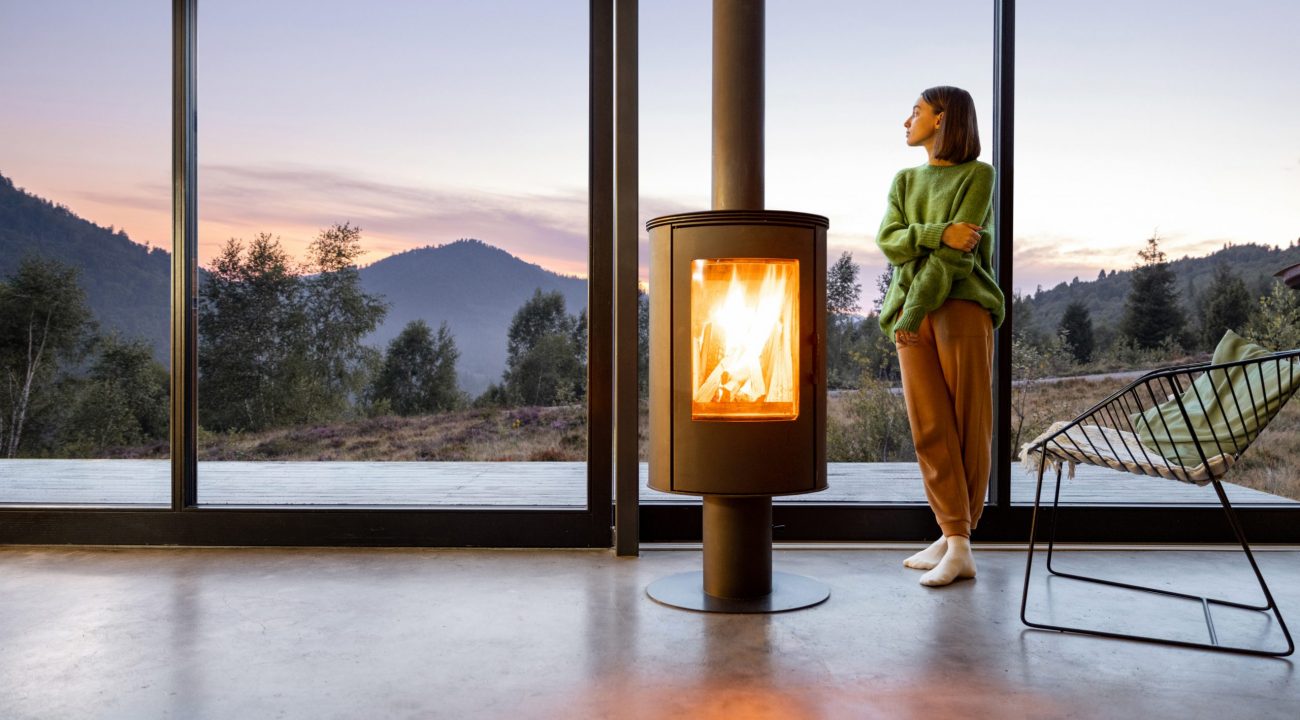 Woman enjoy great view on mountains while sitting near fireplace at modern living room at sunset . Concept of rest in houses or cabins on nature. Solitude in nature and escape from everyday life